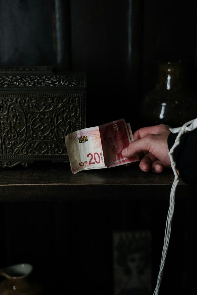 A close-up of a hand holding a 20 NIS currency note against a dark interior background.