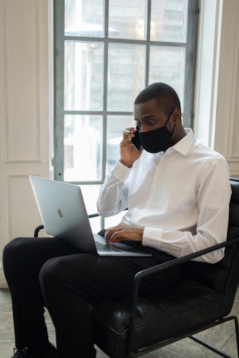 A professional man in a face mask multitasks with laptop and phone indoors.